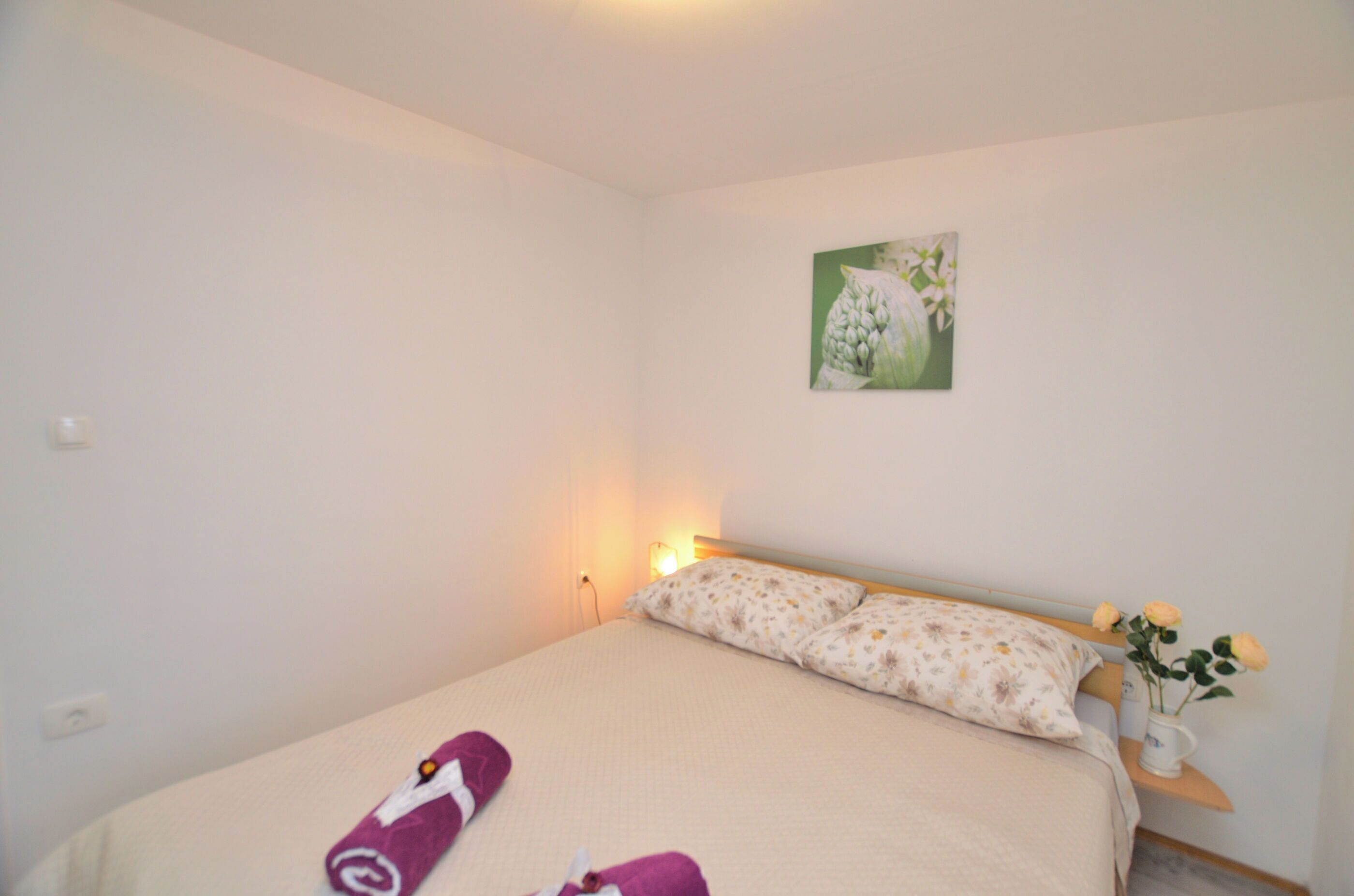 A neatly made bed with floral bedding, a bedside lamp, and a small vase with flowers on a nightstand.