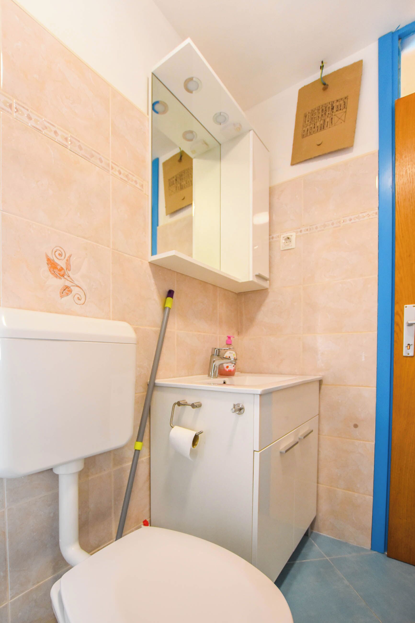 A bathroom with a toilet, sink, mirror, and a mop leaning against the wall.