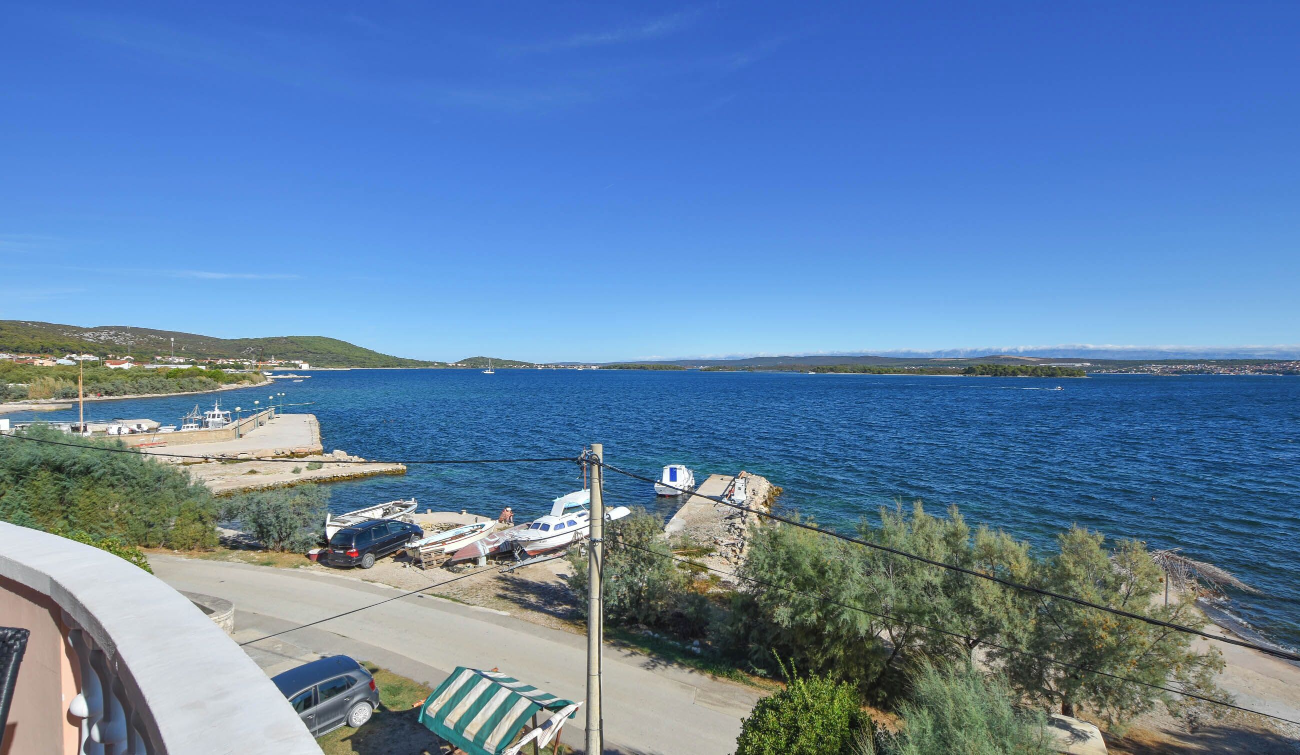A coastal view with boats docked at a marina, a clear blue sky, and a hilly landscape in the distance.