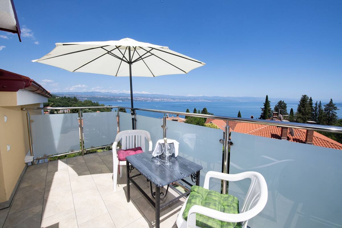 Un balcon avec une table et des chaises, un parasol, et une vue sur la mer et le paysage environnant.