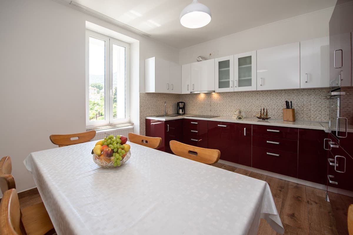 Une cuisine moderne avec des meubles blancs et bordeaux, une table à manger agrémentée d’un panier de fruits, et une vue sur la verdure à l’extérieur.