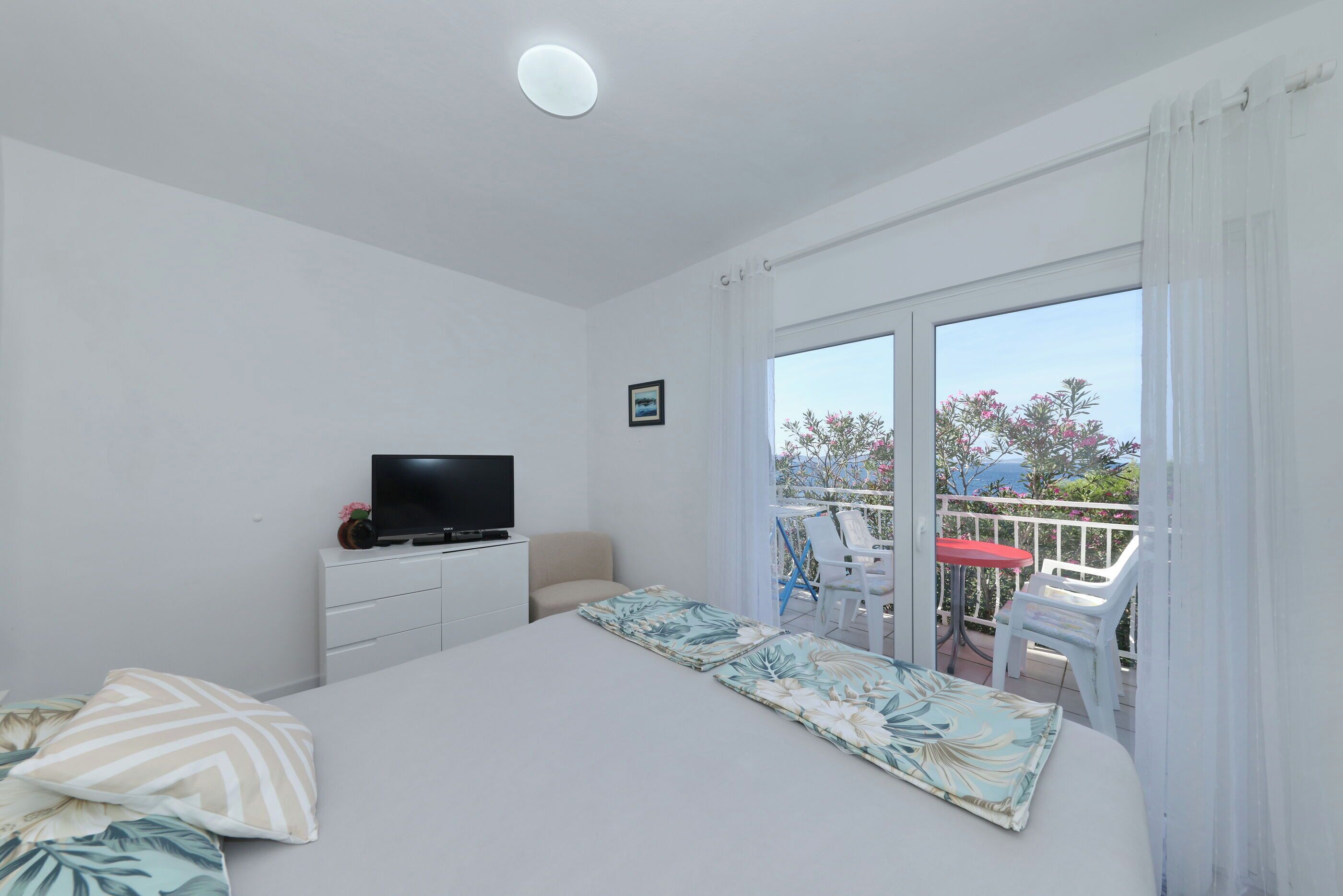 A bedroom with a bed, a TV, a small desk, and a balcony with chairs and a table.