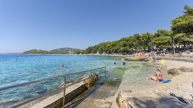 Une plage à l’eau claire, un rivage rocheux, et des gens qui se baignent et se détendent.