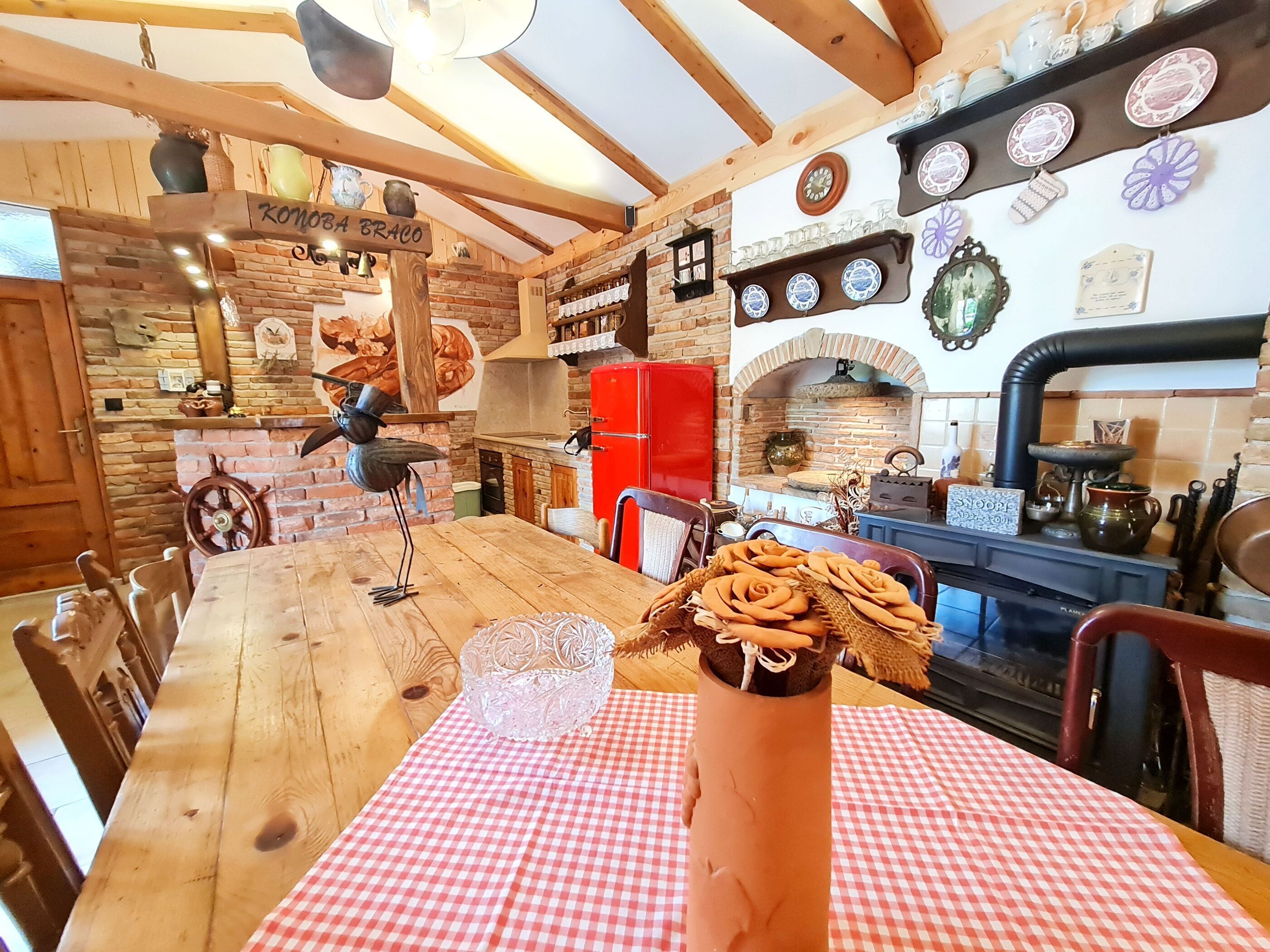 A rustic kitchen with exposed brick walls, wooden beams, a red refrigerator, and a variety of kitchenware displayed on shelves.