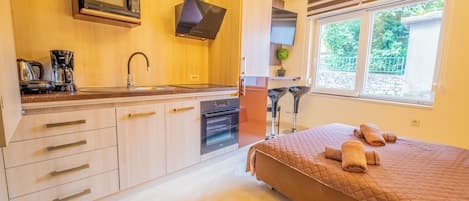 A compact kitchen with a microwave, coffee maker, and oven. A bed with a quilted cover and towels is visible.