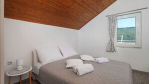 A bedroom with a wooden ceiling, a bed with a beige bedspread, two pillows, a small round table with a lamp, and a window with a view of a landscape. - Apartment Meri (Sibenik)