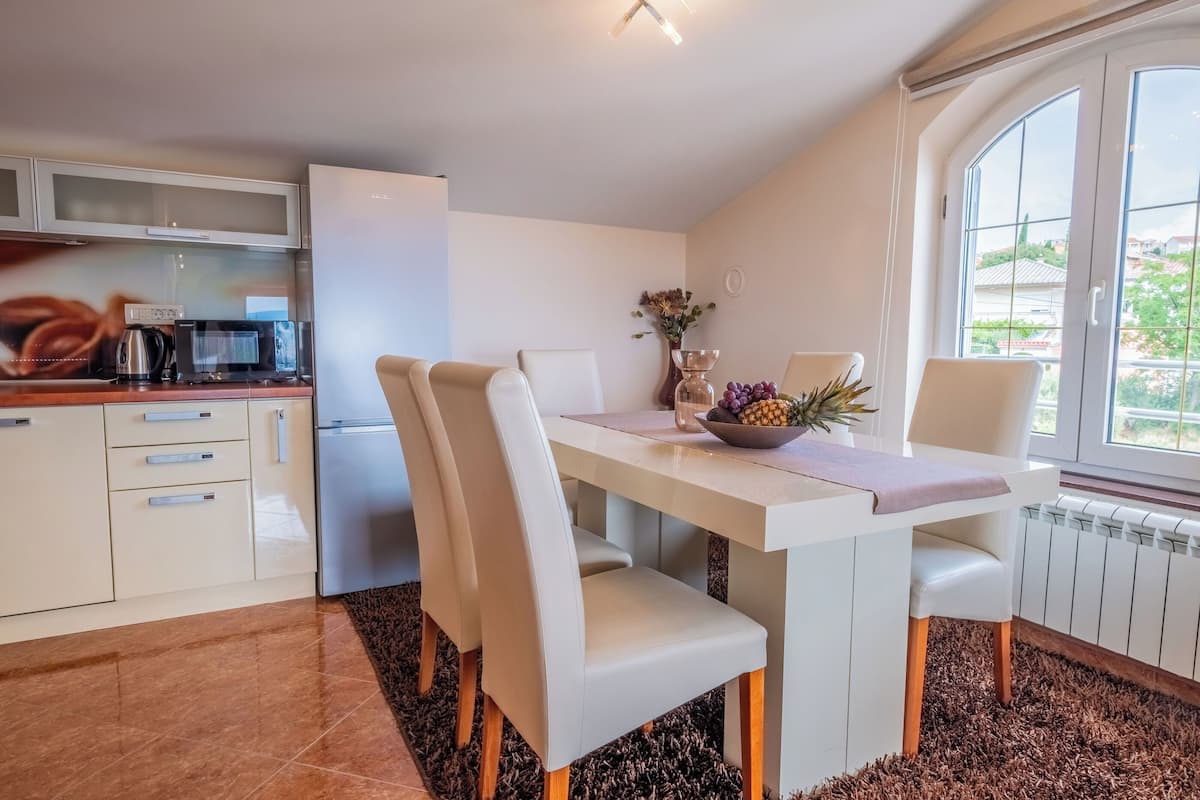 A modern kitchen with white cabinetry, a dining table with chairs, a coffee maker, and a view of the outside through large windows.