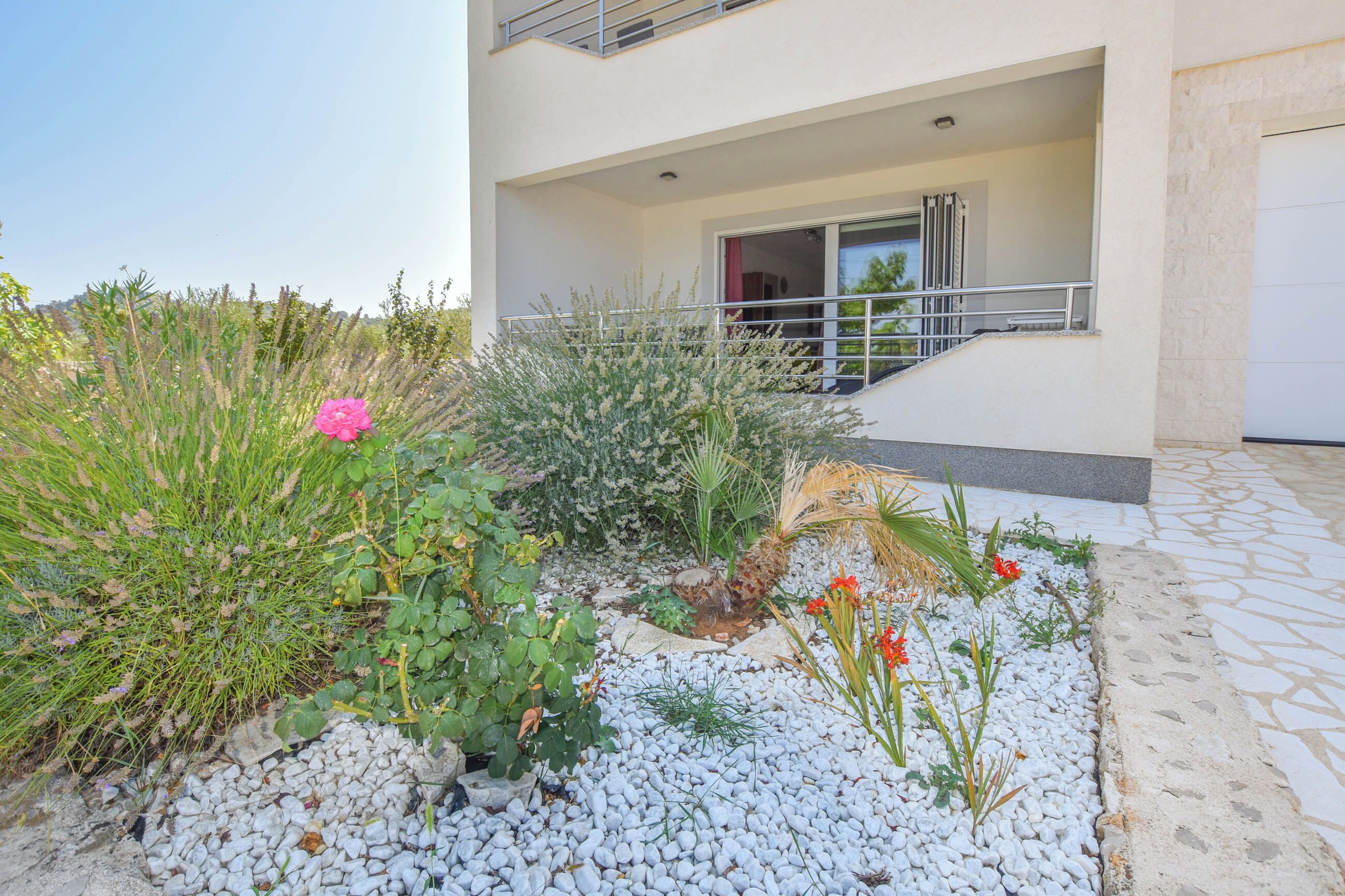 A modern building with a balcony, white stone flooring, and a garden with various plants and flowers.