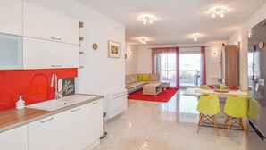 A modern kitchen with white cabinets, a red countertop, and a built-in sink. There is a dining area with a table and chairs, and a living room with a sofa and a red rug. - Apartment Ray of Sunshine (Pasman)