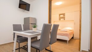 Ein gemütlicher Raum mit Esstisch, Stühlen, einem kleinen Kühlschrank, einer Topfpflanze und einem Bett mit Nachttisch.