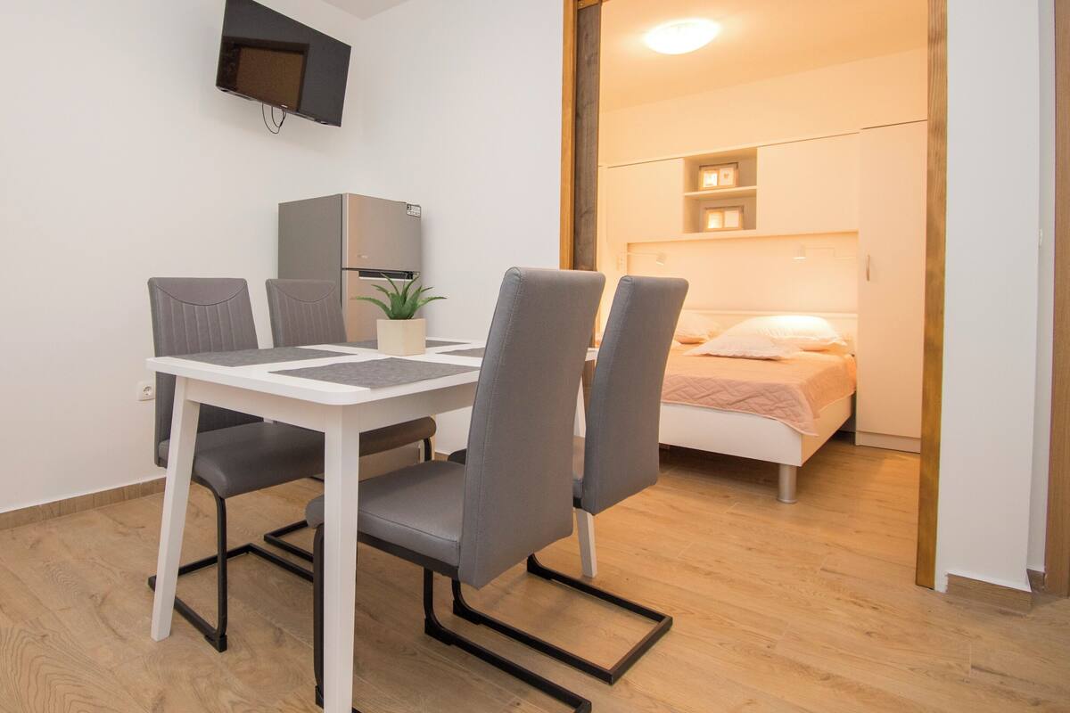 Une pièce de taille modeste comprenant une table à manger, des chaises, un petit réfrigérateur, une plante en pot et un lit avec une table de chevet.