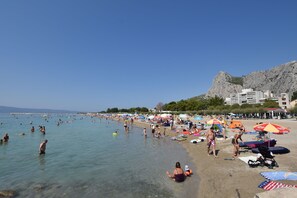 A crowded beach with people swimming and sunbathing, umbrellas, and a rocky hill in the background.