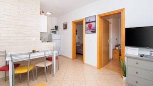 A modern kitchen with white cabinets, a microwave, and a dining area with a table and chairs. There is a TV on a dresser and a plant in the corner. - Apartment Monika (Vodice)