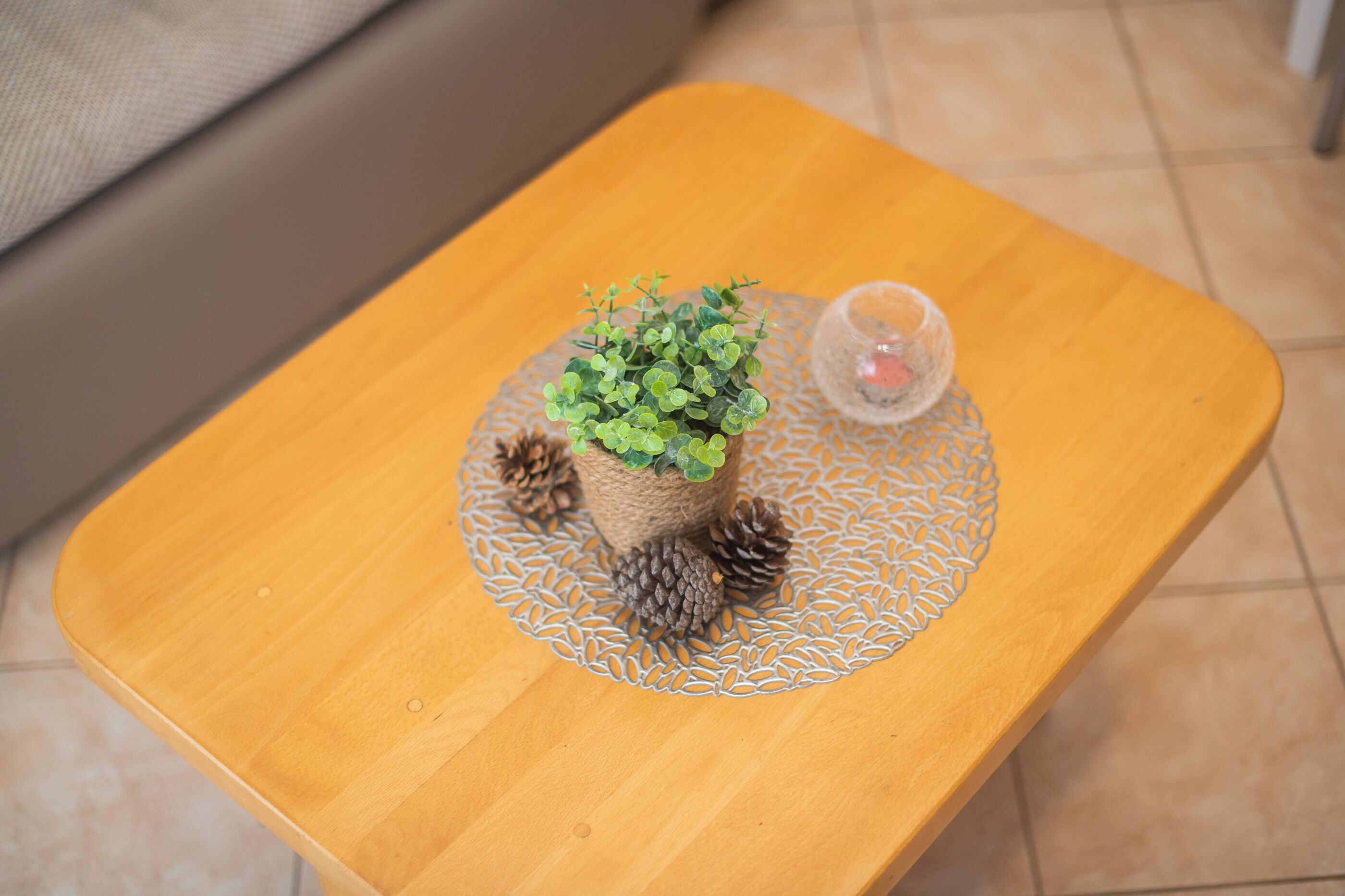 Una pequeña mesa de madera con un portalápices tejido que contiene follaje verde y piñas, y un candelabro de vidrio.
