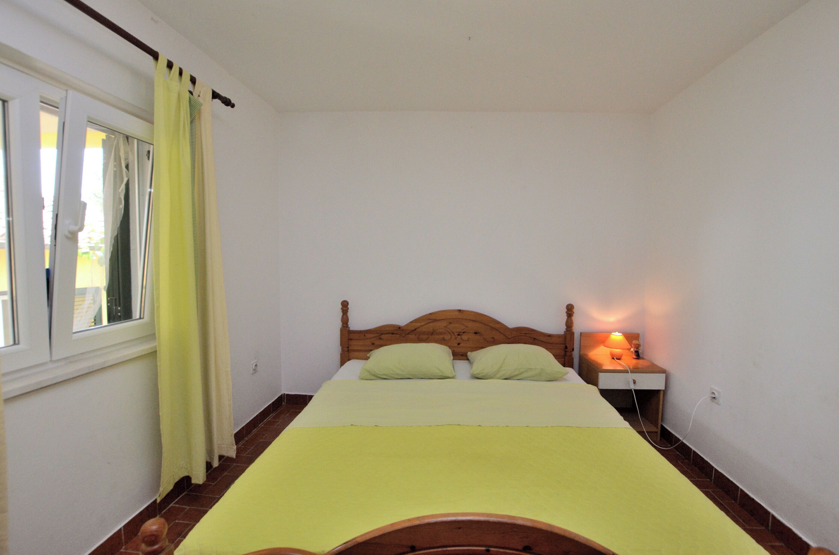 A bedroom with a wooden bed, a nightstand with a lamp, a window with curtains, and a view of the outside.