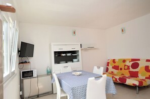 Een lichte kamer met een eettafel, witte stoelen, een bank, een televisie en een magnetron.