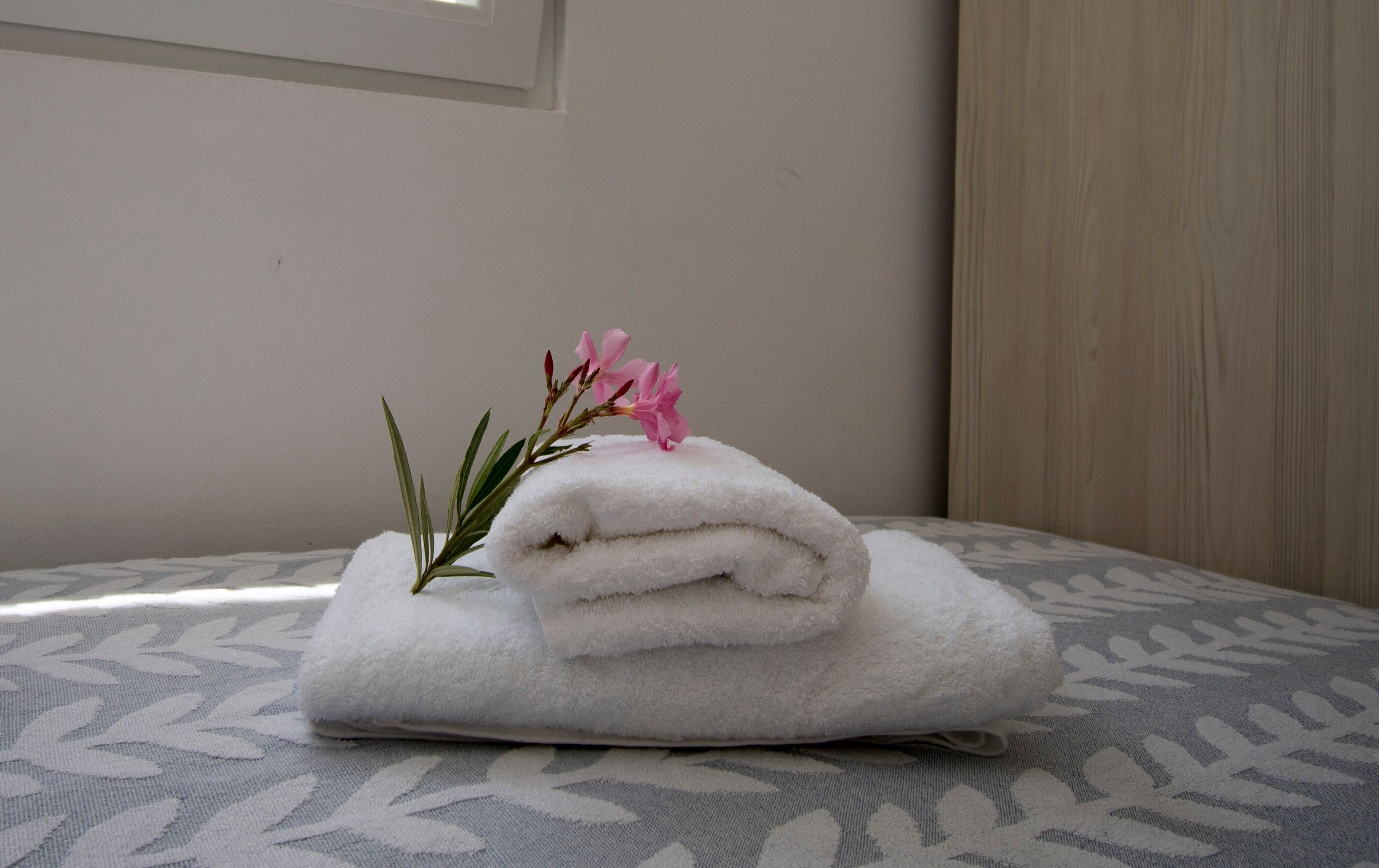 A neatly folded white towel with a sprig of pink flowers on a patterned gray and white bedspread.
