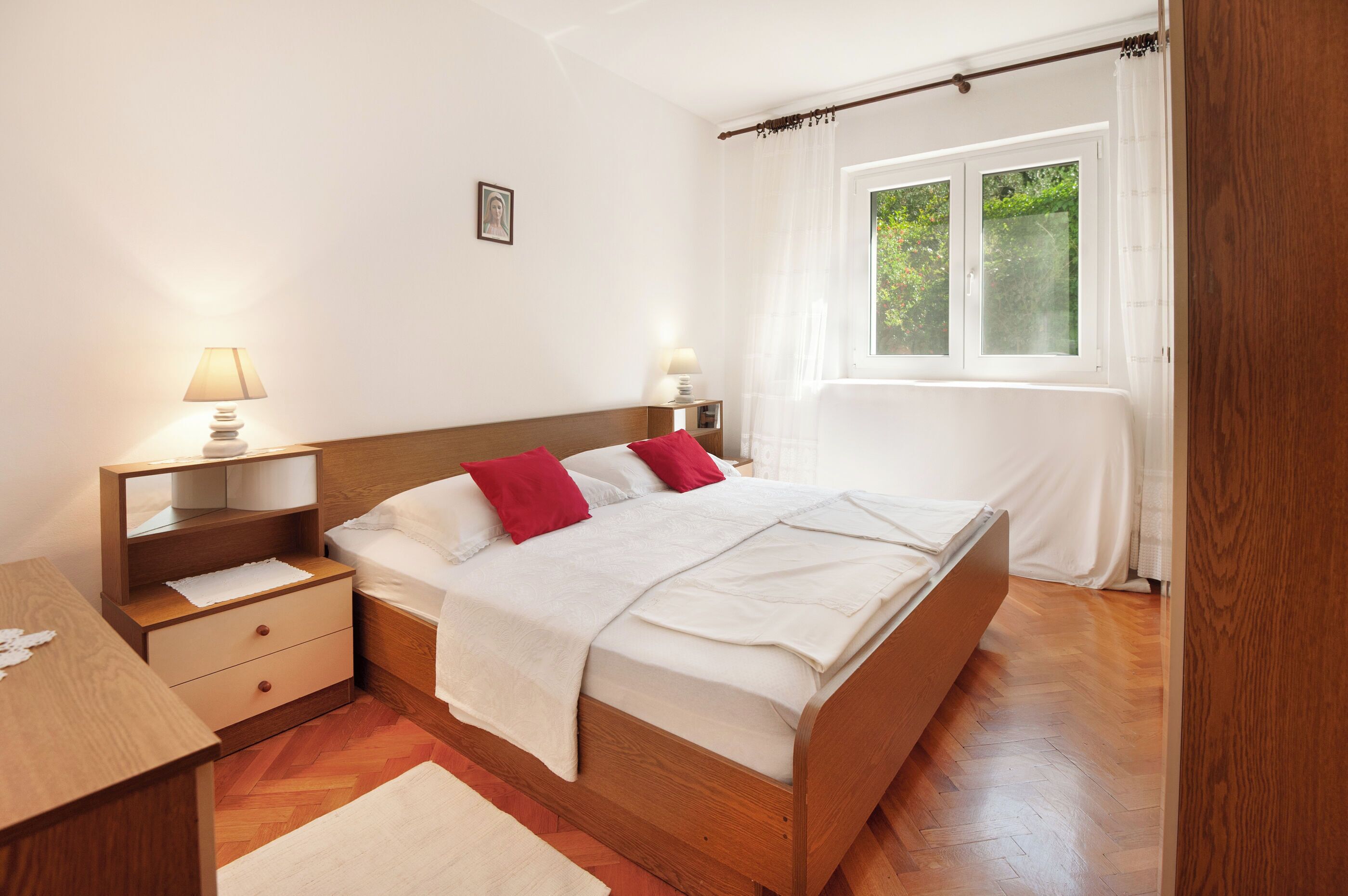A bedroom with a wooden bed, white bedding, a nightstand with a lamp, and a window with curtains.