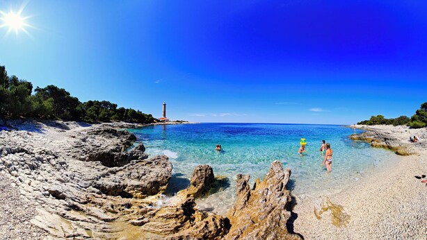 A beach with clear water, rocky shore, and a lighthouse in the distance. - Apartments Veli Rat (Sali)