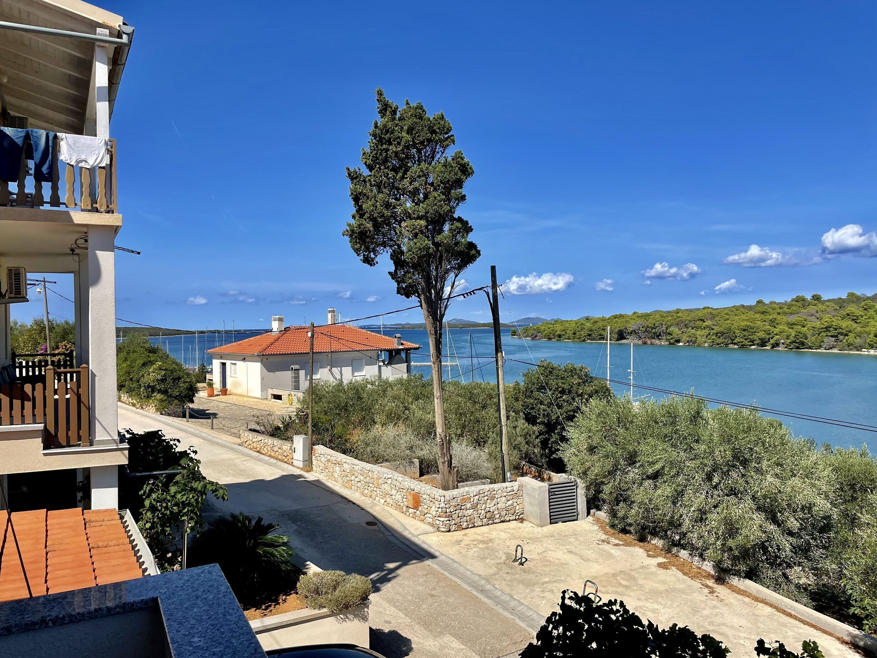A coastal property with a balcony overlooking a clear blue sea, a stone wall, and a small building with a red roof.