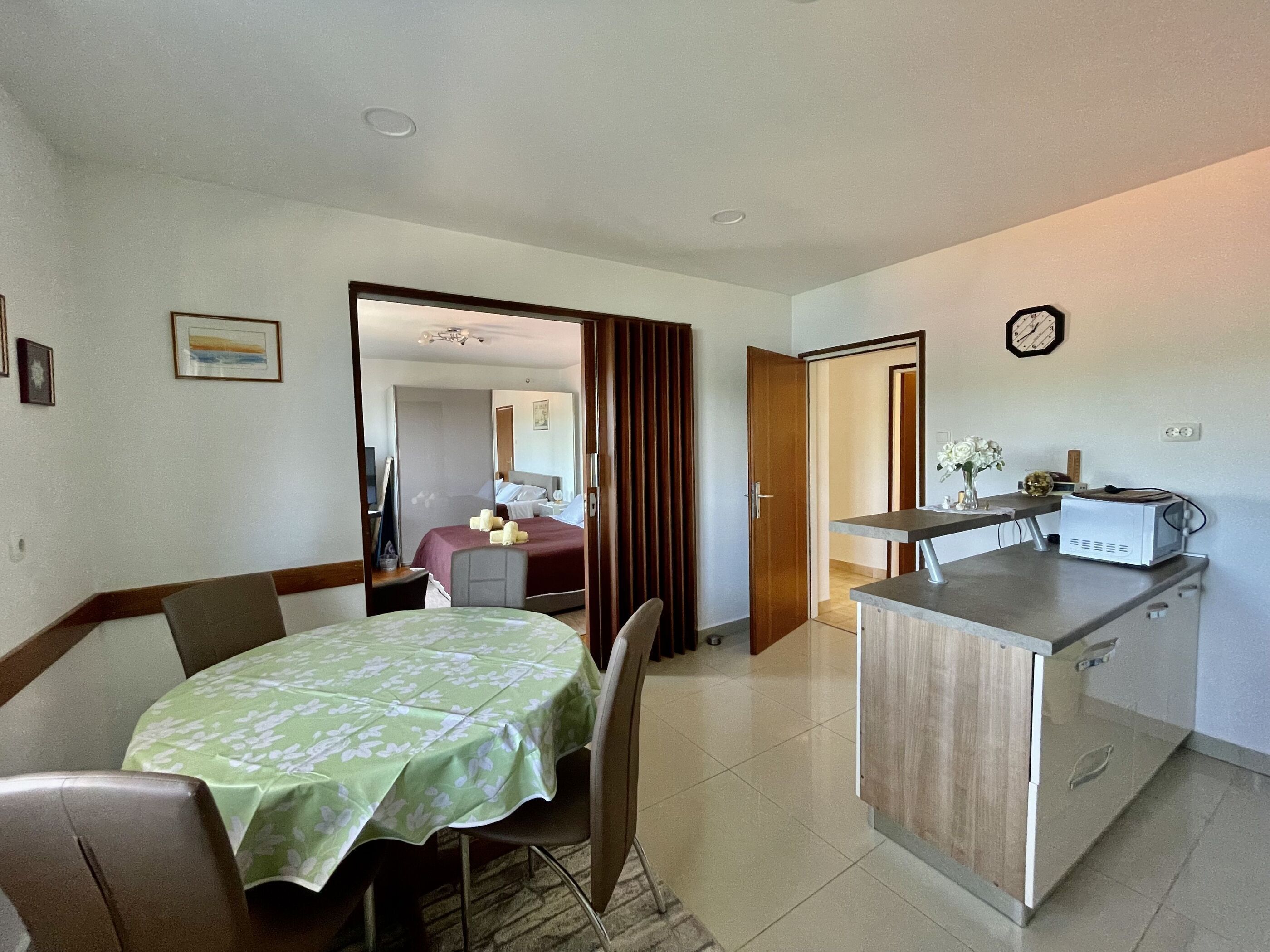 A hotel room with a dining table, chairs, a kitchen area, and a bed in the background.