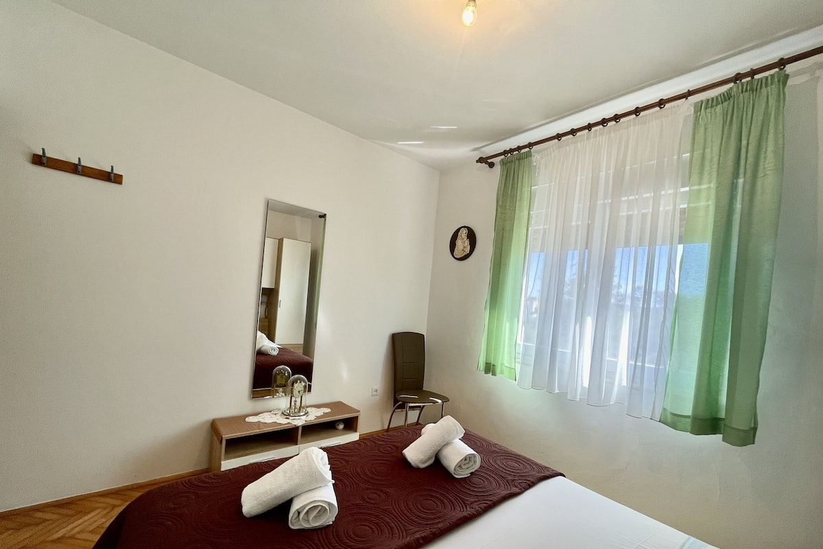 A hotel room with a bed, a bedside table, a mirror, a chair, and a window with sheer curtains.