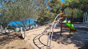 A playground with a climbing structure and a slide, surrounded by trees and a view of the sea. - Apartments On the Beach (Sucuraj)