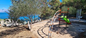 A playground with a climbing structure and a slide, surrounded by trees and a view of the sea. - Apartments On the Beach (Sucuraj)