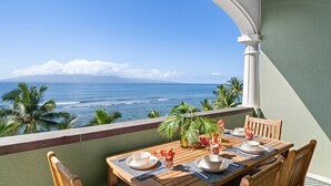 Un balcón con mesa de madera dispuesta para comer, con vistas a un paisaje marino con palmeras y cielo despejado.