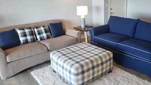Una sala de estar con un sofá beige, un sofá azul y un otoman tejido. Hay una mesita con una lámpara entre los sofás.