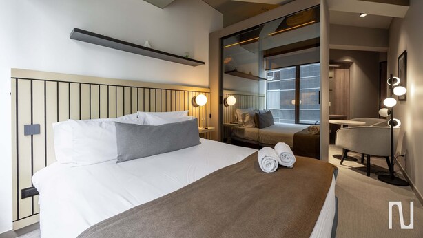 Quarto de hotel moderno com uma cama grande, mesas de cabeceira, um espelho e um espaço de estar com uma luminária.