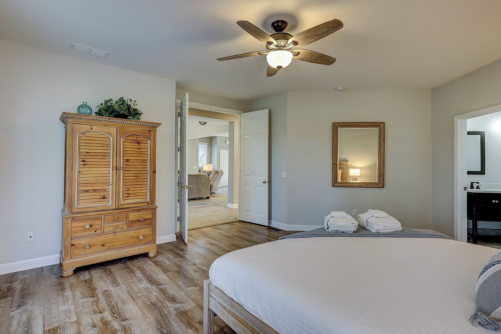 A bedroom with a bed, a wooden wardrobe, a ceiling fan, a mirror, and a door leading to another room.