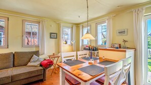 Una sala de estar acogedora con un sofá, una mesa de comedor con sillas, una pequeña cocina y ventanas grandes con cortinas.