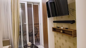Un salón moderno con un televisor de pantalla plana montado en la pared, un puerta corrediza de cristal que da a un balcón y una pequeña mesa redonda con dos tazas blancas.