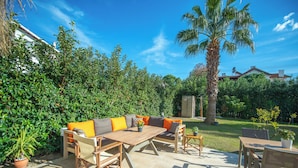 An outdoor patio with a wooden table and chairs, a sofa with orange cushions, and a palm tree. - Kalender Pansiyon Fethiye (Fethiye)