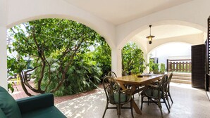 Un espace repas avec une table et des chaises en bois, entouré d’arches blanches et d’une végétation luxuriante.