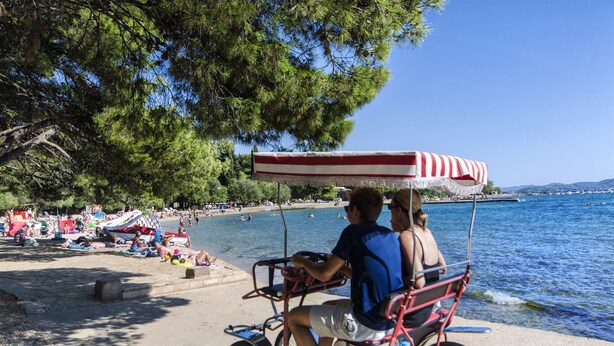 Une scène de plage avec un parasol à rayures rouges et blanches, une zone de plage où se trouvent des personnes, et une étendue d’eau.