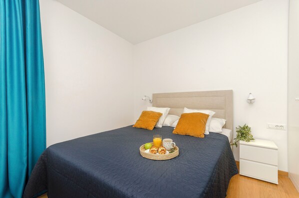 Un lit bien fait, recouvert d’une couvre-lit bleu foncé, avec deux oreillers blancs et un seul oreiller orange. Une petite table de chevet porte une plante et une lampe blanche. Un plateau contenant des éléments de petit-déjeuner, notamment des fruits et une boisson, est posé sur le lit.