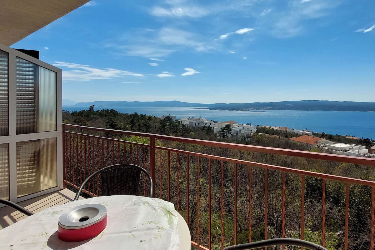 A balcony with a table and chairs, overlooking a coastal view with buildings and mountains in the distance.