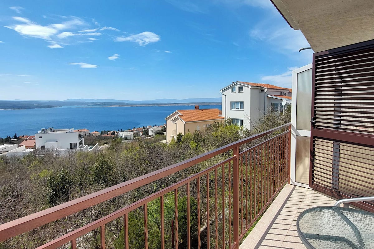 A balcony with a view of the sea, a white building with a red roof, and a glass table.