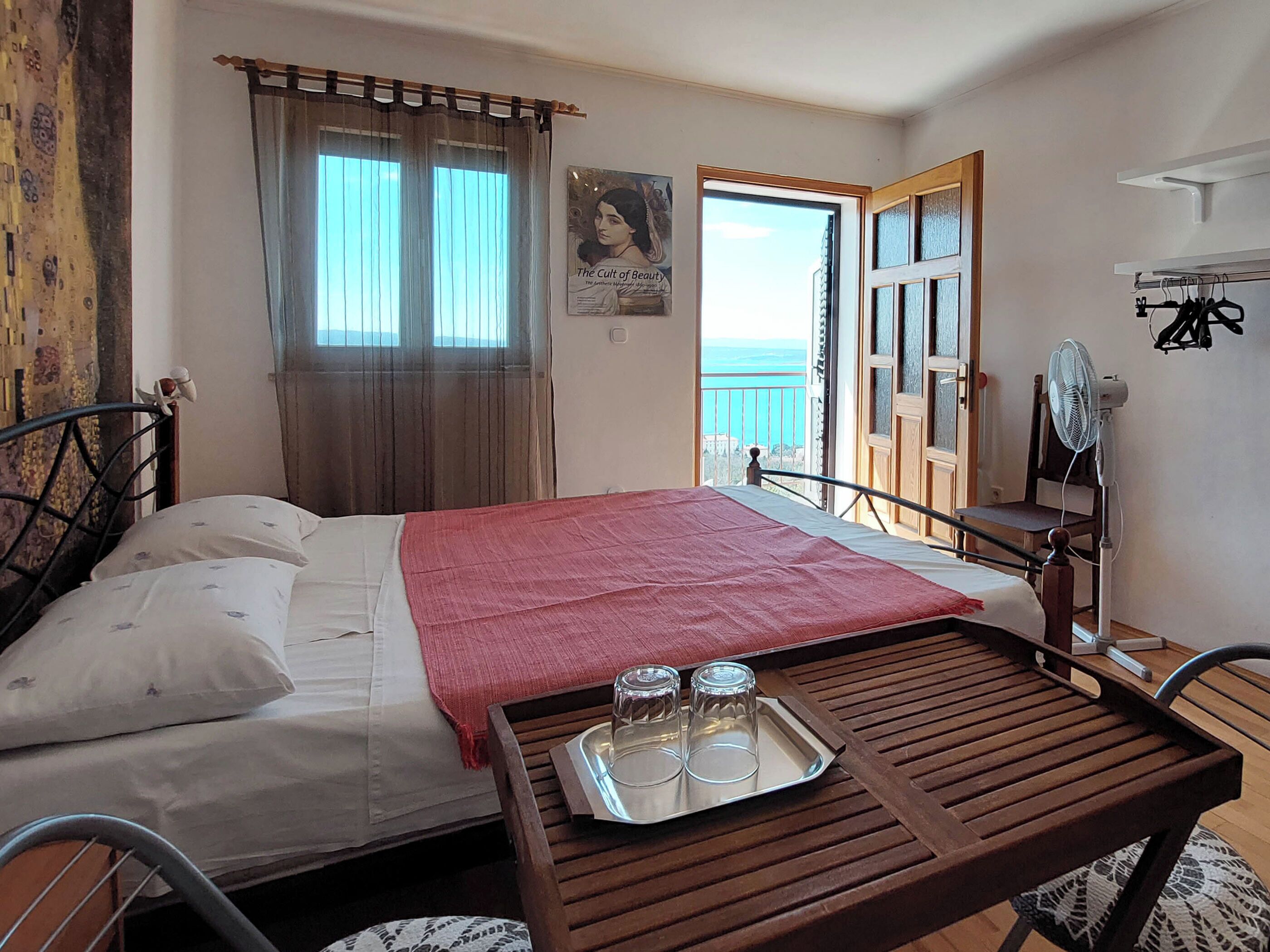 Un dormitorio con cama, una mesa de madera con vasos y una bandeja, una ventana con cortinas, un cartel en la pared y un balcón con vistas.