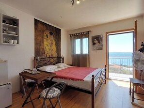 A bedroom with a bed, a small table with a chair, a ceiling fan, a window with curtains, and a painting on the wall. - Apartments Dara (Crikvenica)