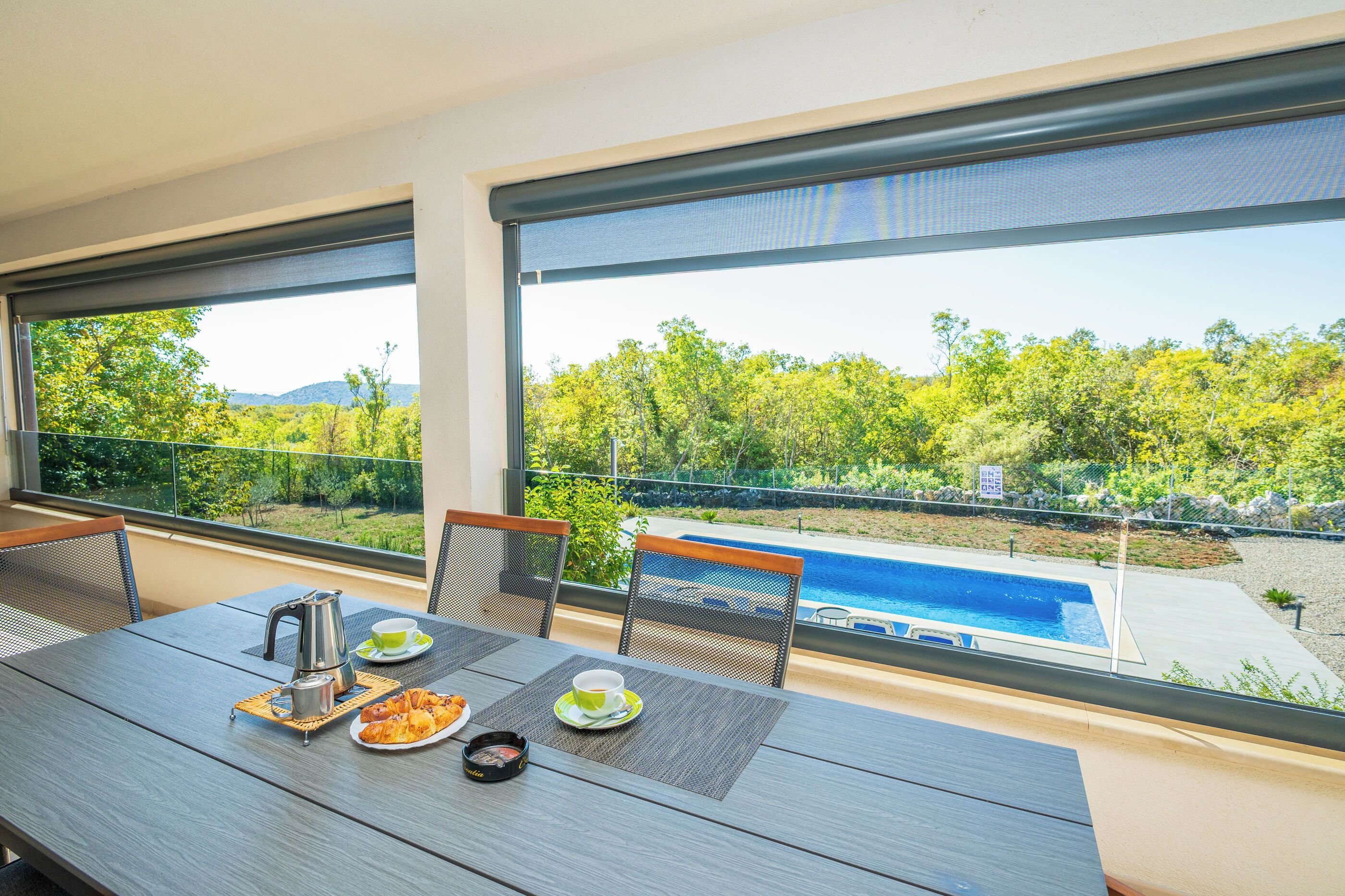 Comedor con una mesa para dos, vistas a una piscina y abundante vegetación.