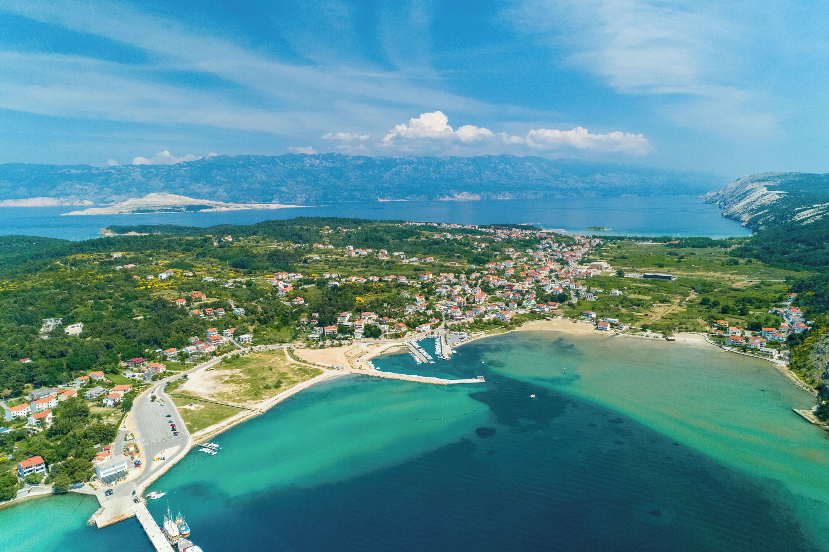 An aerial view of a coastal town with a marina, residential areas, and mountains in the background.