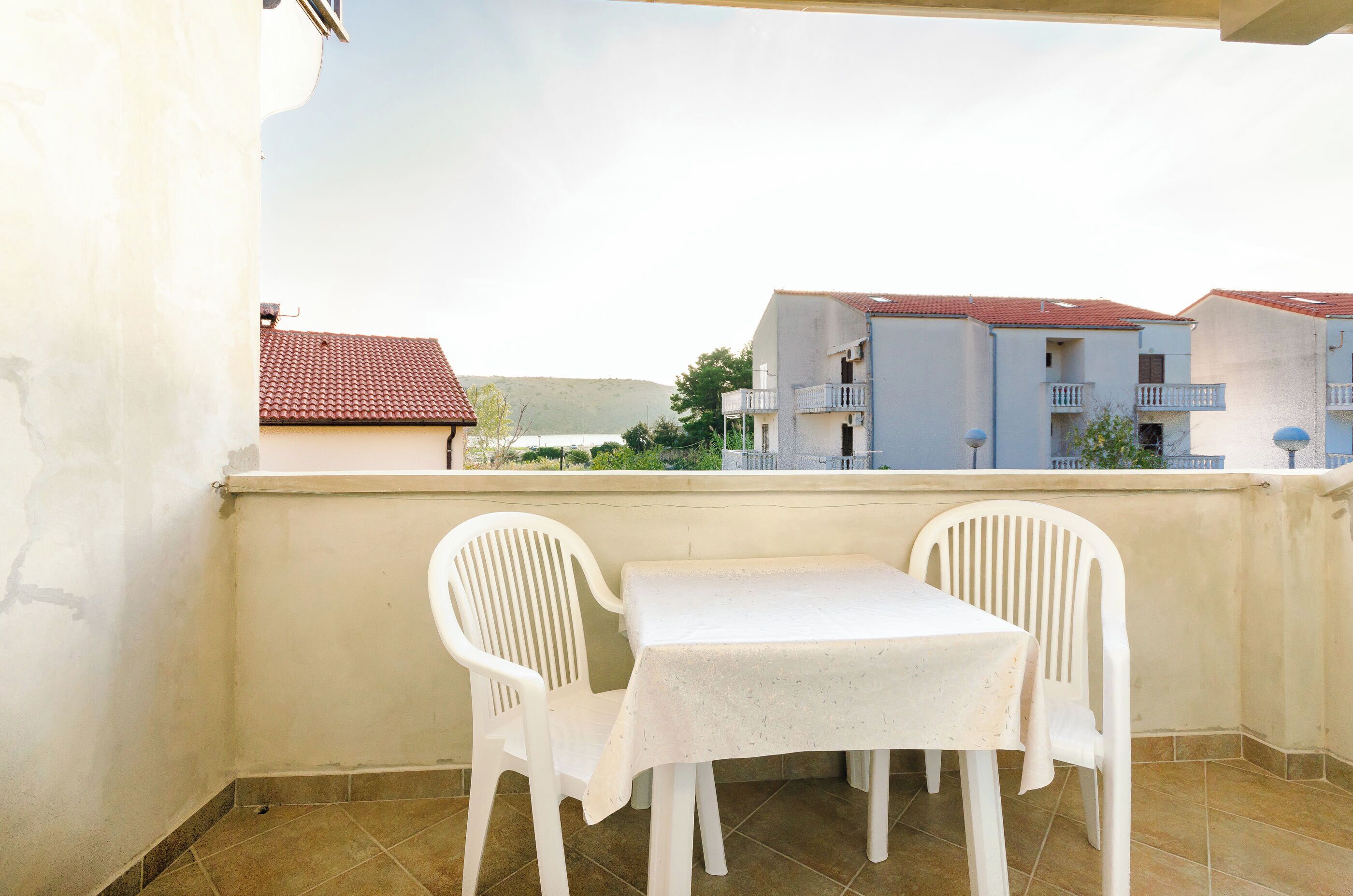 A balcony with a white table and two white chairs, overlooking a residential area with buildings and a hill.