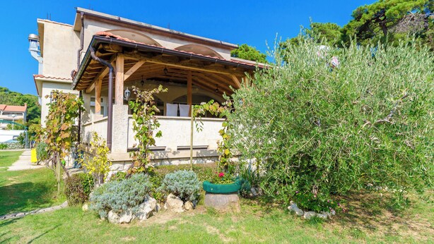 A house with a wooden porch, a garden with various plants, and a clear blue sky. - Apartments Josip S (Lopar)