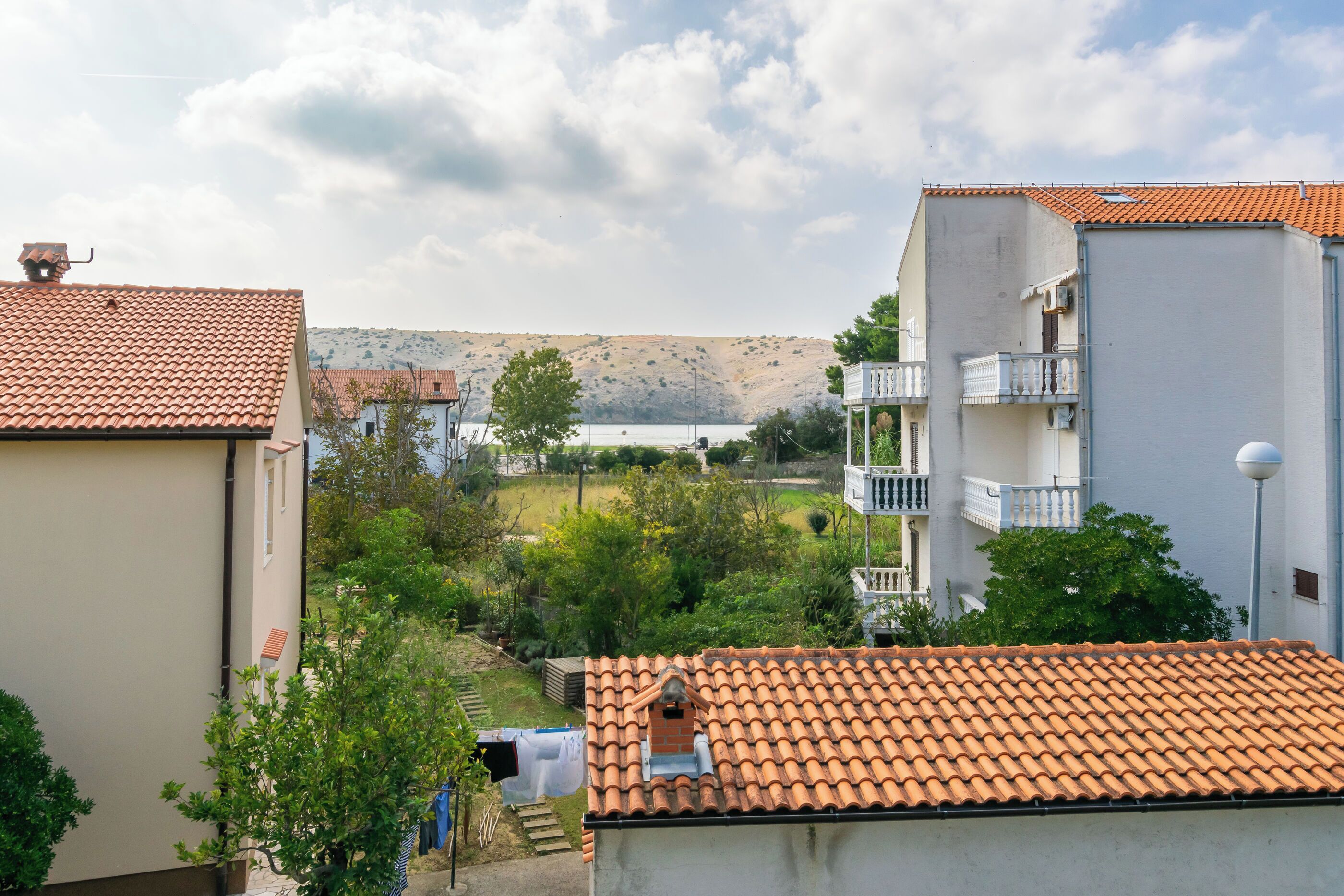 A residential area with terracotta-tiled roofs, a hillside in the background, and a white building with a balcony.