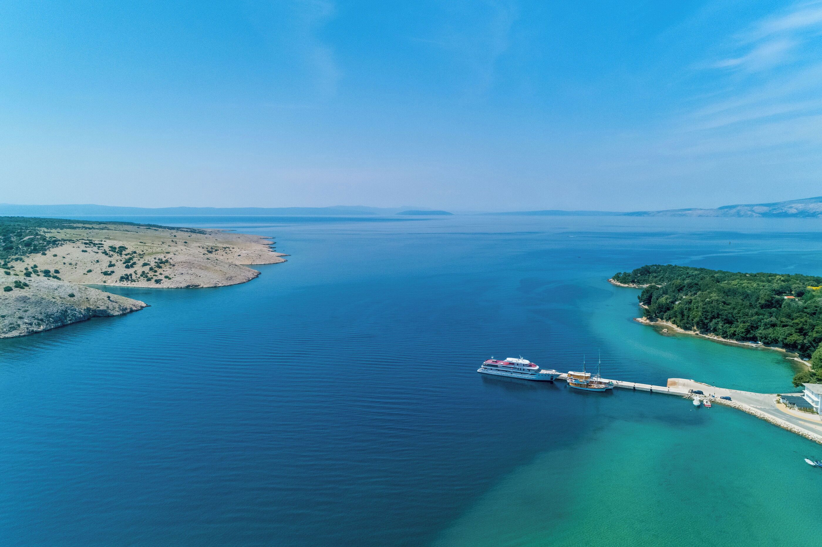 An aerial view of a coastal area with a pier extending into a calm sea, surrounded by land with sparse vegetation and a clear sky.