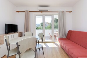Ein modernes Wohnzimmer mit einem roten Sofa, einem Esstisch mit Stühlen, einem Fernseher auf einer Holzkommode und großen Schiebegläsern, die zu einem Außenbereich führen.