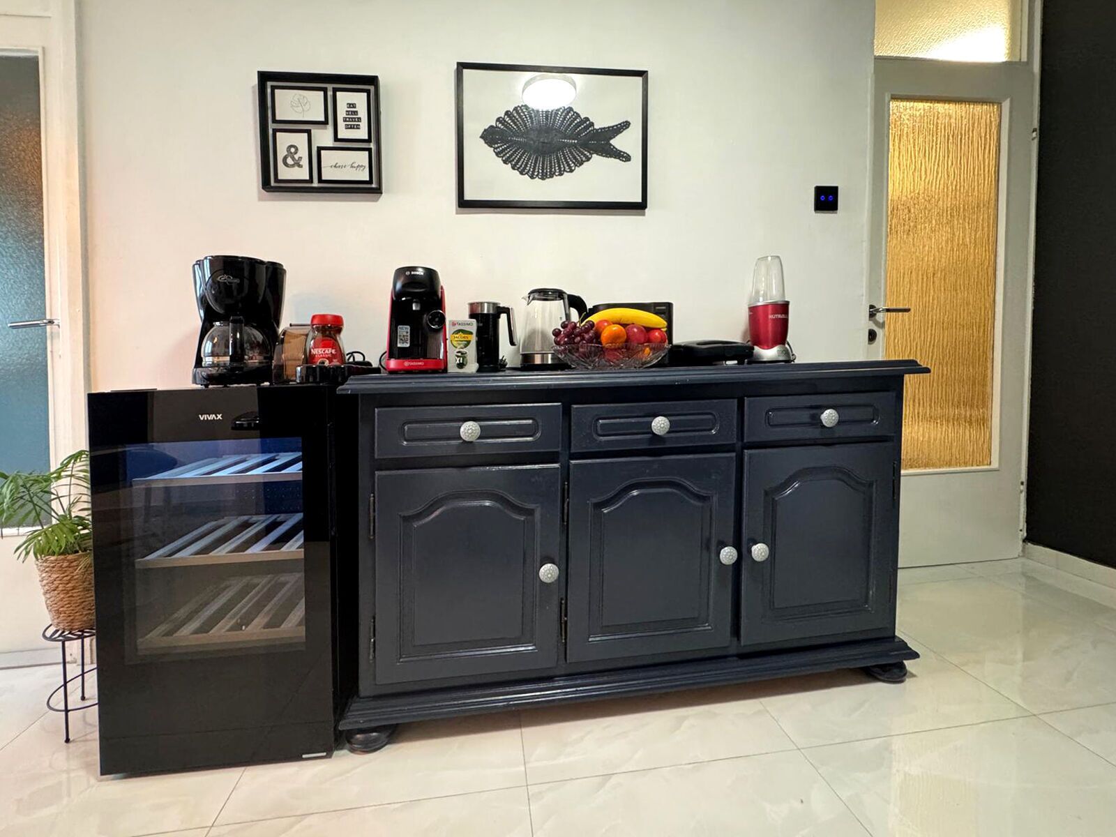 A kitchen with a black refrigerator, a coffee maker, a blender, and a fruit bowl on the counter.